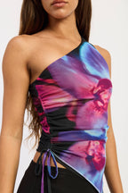 A close-up of a model wearing a one-shoulder ruched top with a vibrant purple, pink, and blue abstract floral print over a black mesh base.