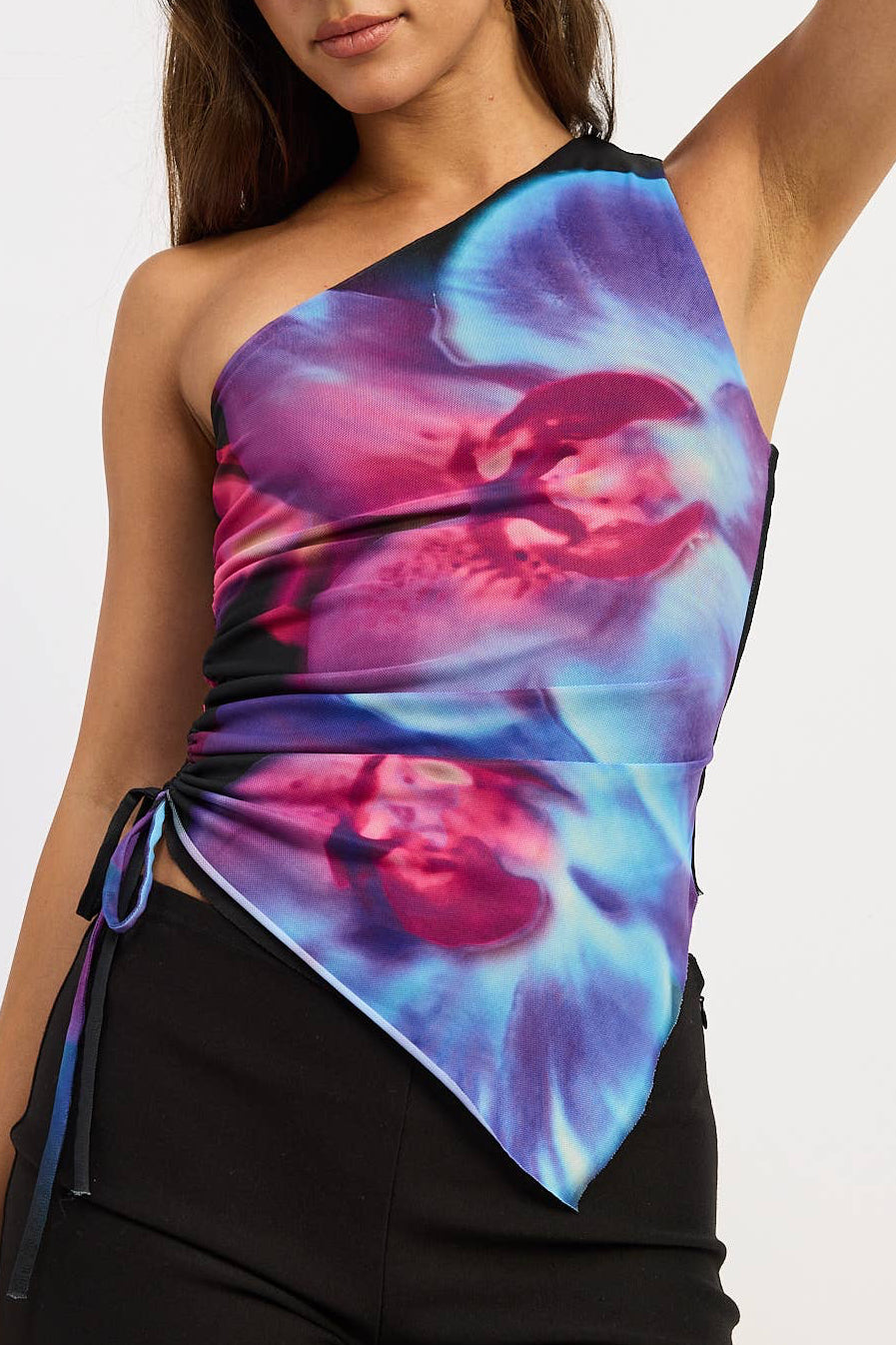 A woman modeling a one-shoulder wrap top with a vibrant blue, purple, and pink abstract print, paired with black shorts, against a plain light gray background.
