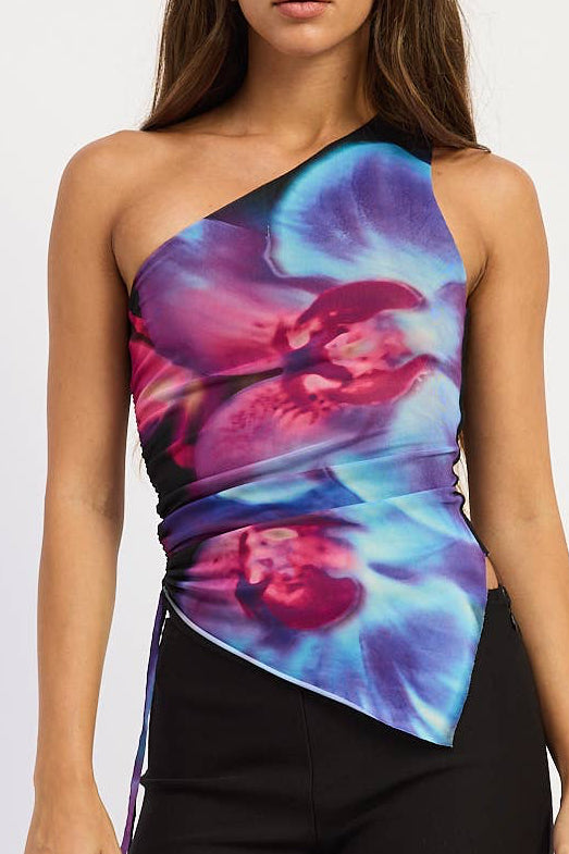 A woman modeling a one-shoulder wrap top with a vibrant blue, purple, and pink floral print, paired with black shorts and black knee-high boots, against a plain white studio background.