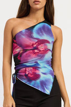 A woman modeling a one-shoulder wrap top with a vibrant blue, purple, and pink floral print, paired with black shorts and black knee-high boots, against a plain white studio background.