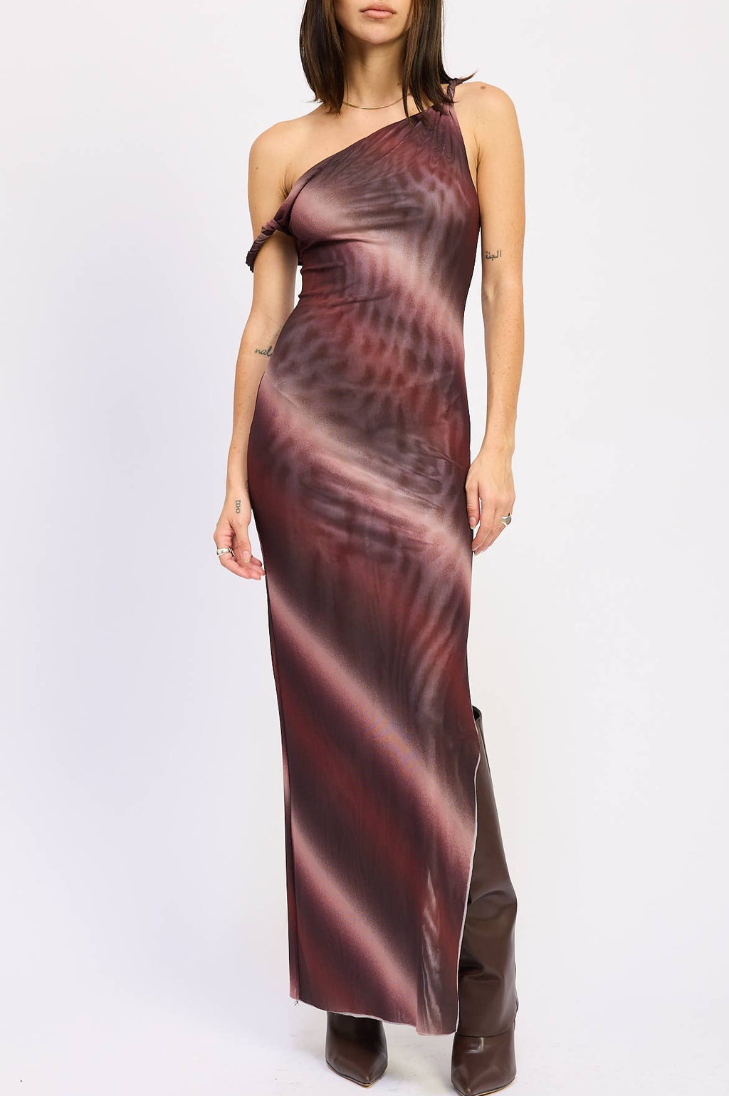A woman modeling a one-shoulder mesh maxi dress with a dark red and black abstract pattern, paired with dark brown boots, against a plain white studio background.