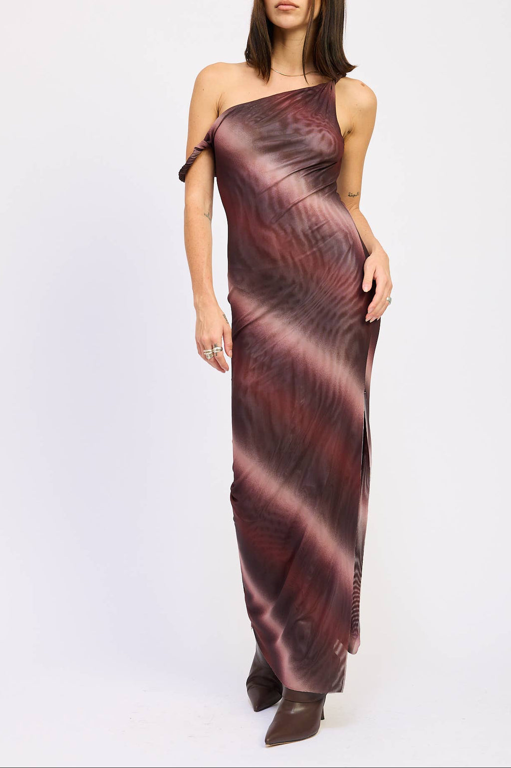 A woman modeling a one-shoulder maxi dress with a dark abstract pattern in deep burgundy, brown, and black, paired with dark brown pointed-toe boots, against a plain white studio background.