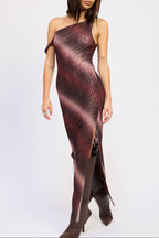A woman modeling a one-shoulder, floor-length dress with a dark abstract print in deep burgundy, brown, and black tones, paired with dark brown pointed-toe boots, against a plain light gray studio background.