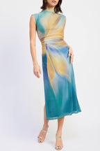 A woman modeling a sleeveless midi dress with a high mock neck and a flowing abstract print in teal blue, gold, and cream, paired with clear high-heeled sandals against a light gray studio background.