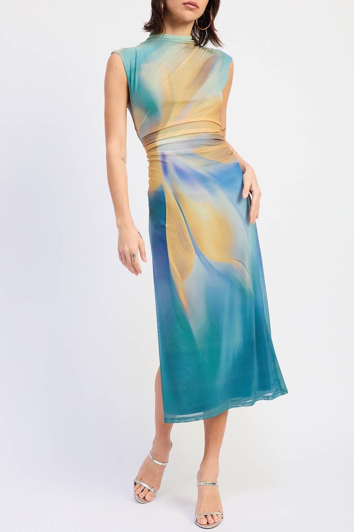 A woman modeling a sleeveless midi dress with a high mock neck and a flowing abstract print in teal blue, gold, and cream, paired with clear high-heeled sandals against a light gray studio background.