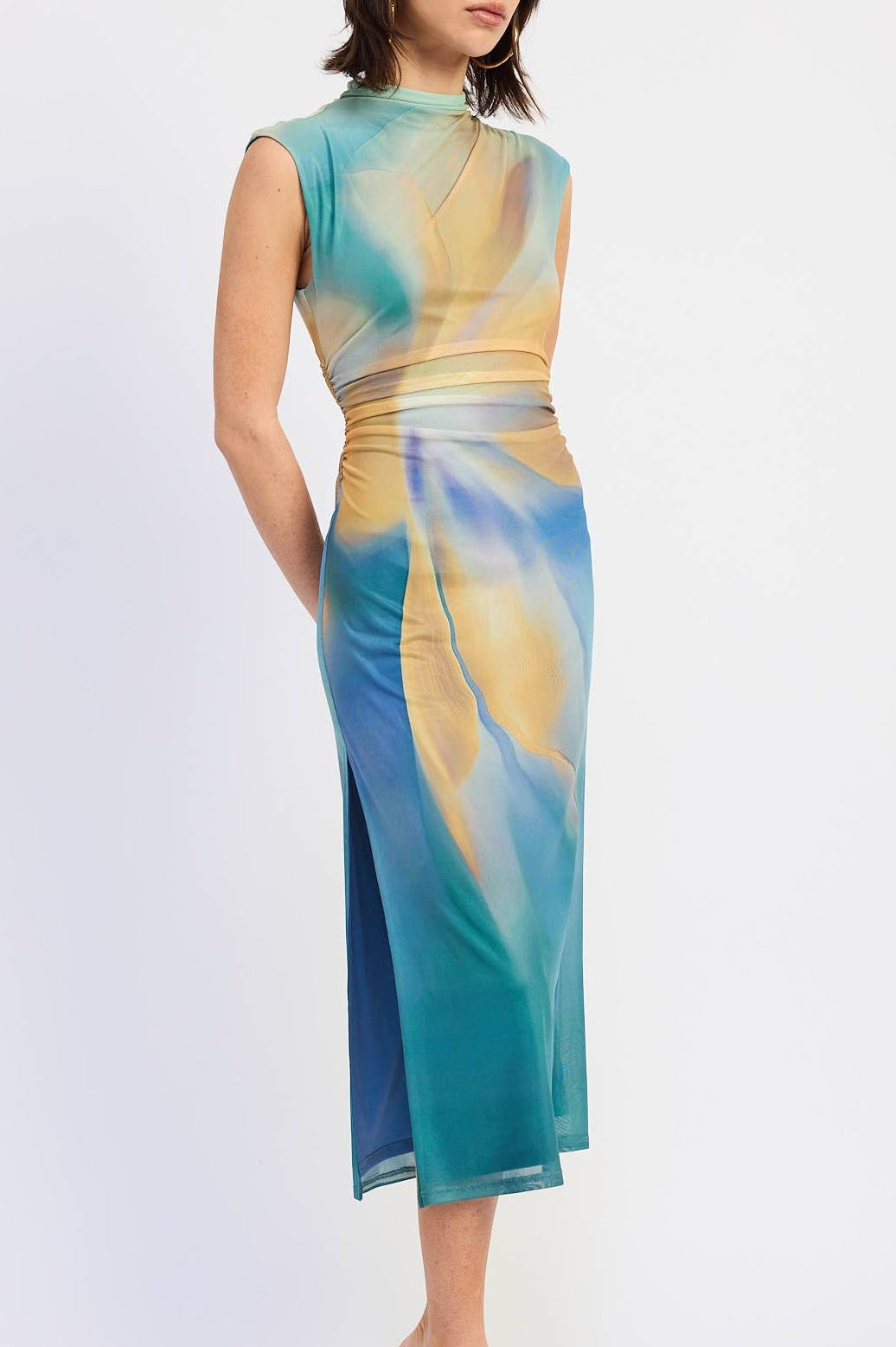 A woman modeling a sleeveless midi dress with a high mock neck and side slit, featuring an abstract watercolor-like print in teal, blue, yellow, and beige on a light gray studio background.