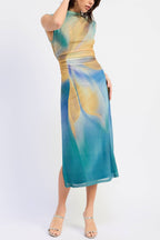 A woman modeling a sleeveless midi dress with an abstract watercolor-like print in teal, blue, and golden yellow, paired with clear strap high-heeled sandals against a plain white studio background.