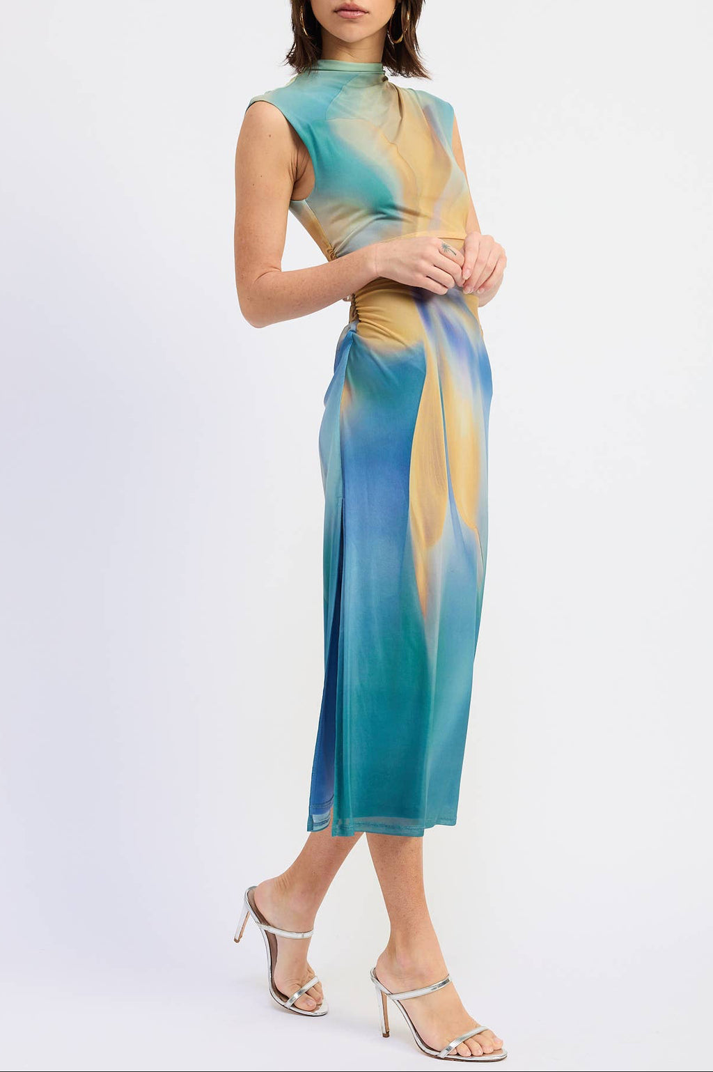 A woman modeling a sleeveless midi dress with a high mock neck, featuring an abstract watercolor-like print in teal blue, turquoise, and mustard yellow, paired with silver high-heeled sandals against a plain white studio background.