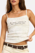 A woman modeling a white camisole tank top with black gothic-style text reading 'It's lost, return to the nearest bar'. She is wearing cream pants, a brown belt, and black sunglasses against a plain white background.