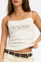 A woman modeling a white camisole tank top with black gothic-style text reading 'It's lost, return to the nearest bar'. She is wearing cream pants, a brown belt, and black sunglasses against a plain white background.