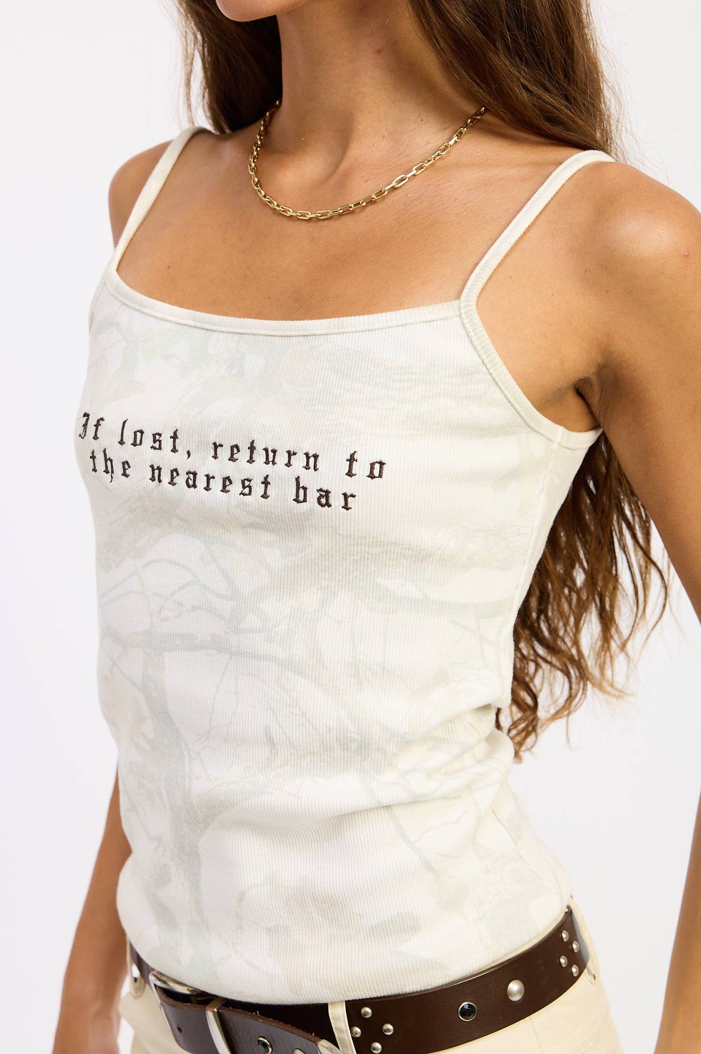 A woman wearing a white camisole tank top with black text that reads 'If lost, return to the nearest bar' across the chest, paired with a brown belt and cream pants against a plain white background.
