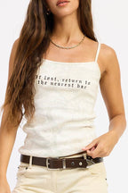 A woman modeling a white spaghetti-strap tank top with black text, cream-colored flared jeans, a brown belt, and black platform shoes against a plain white background.