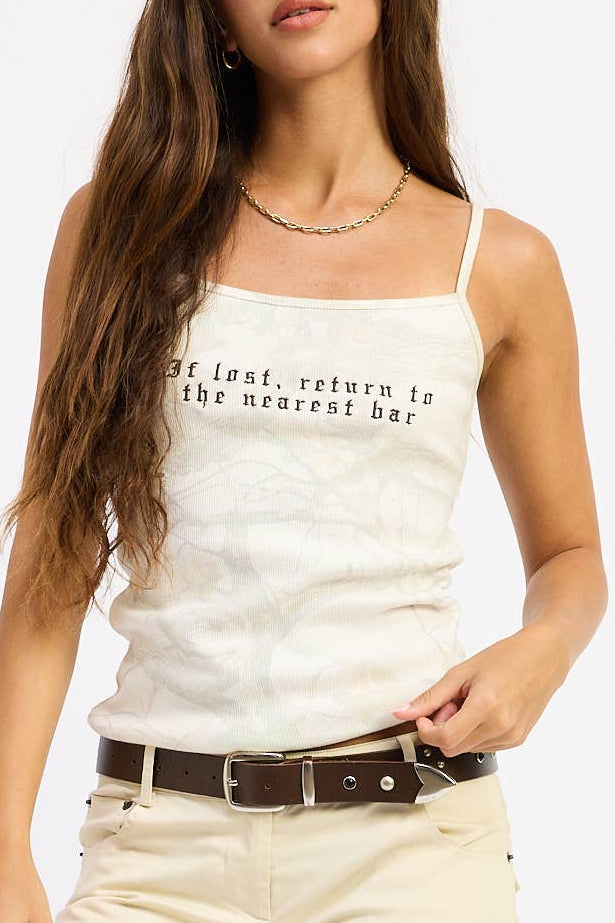 A woman modeling a white spaghetti-strap tank top with black text, cream-colored flared jeans, a brown belt, and black platform shoes against a plain white background.