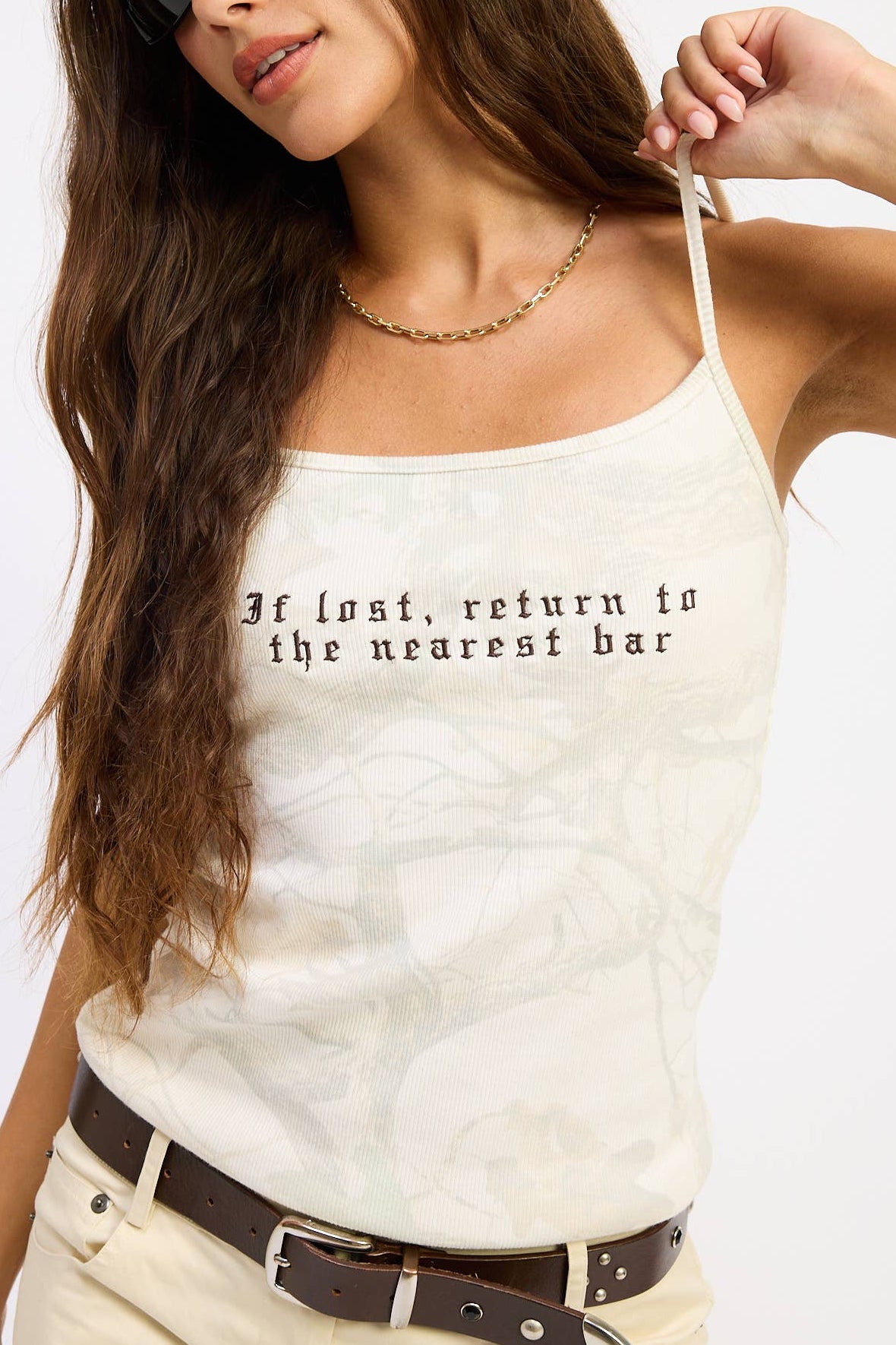 A woman wearing a cream-colored spaghetti-strap tank top with black gothic text that reads 'If lost, return to the nearest bar'. She is also wearing beige pants, a brown belt, and black sunglasses against a plain white background.
