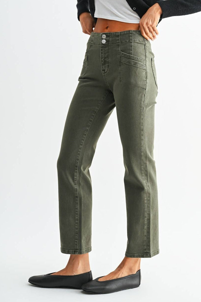 A woman modeling a pair of olive green straight-leg jeans, styled with a black cropped cardigan, a white crop top, and black flats, against a light gray studio background.