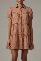 A woman stands barefoot against a plain light gray studio background wearing a short puff-sleeve tiered dress with a small-scale check pattern in muted red and beige.