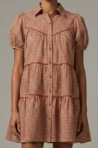 A model wearing a short-sleeve tiered shirt dress with a brown and beige gingham pattern and matching brown trim.