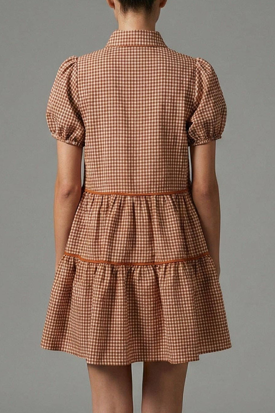 A woman stands with her back to the camera wearing a short puff-sleeve dress with a brown and beige gingham pattern and a tiered skirt, photographed against a neutral gray studio background.