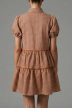 A woman stands with her back to the camera wearing a short puff-sleeve dress with a brown and beige gingham pattern and a tiered skirt, photographed against a neutral gray studio background.
