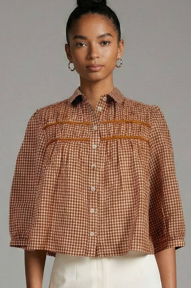 A woman wearing a cropped brown and beige gingham button-up shirt with horizontal orange-brown stripe accents across the chest, paired with white pants, against a solid gray studio background.