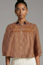 A woman wearing a cropped brown and beige gingham button-up shirt with horizontal orange-brown stripe accents across the chest, paired with white pants, against a solid gray studio background.