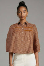 A model stands against a neutral gray studio background wearing a cropped, short-sleeved button-up shirt with a brown and white gingham pattern and pleated details, paired with high-waisted, wide-leg white trousers.