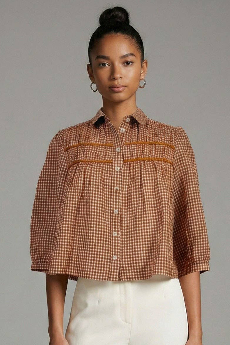 A model stands against a neutral gray studio background wearing a cropped, short-sleeved button-up shirt with a brown and white gingham pattern and pleated details, paired with high-waisted, wide-leg white trousers.