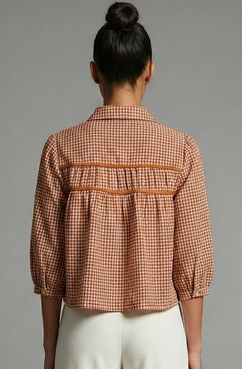 A woman viewed from behind wearing a rust-orange and white gingham cropped blouse with pleated back detail and white wide-leg trousers, standing against a neutral gray studio background.