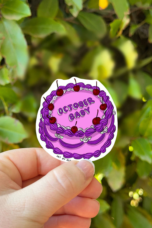 Hand holding a sticker of a purple cake with red berries and 'October Baby' text against a green leafy background.