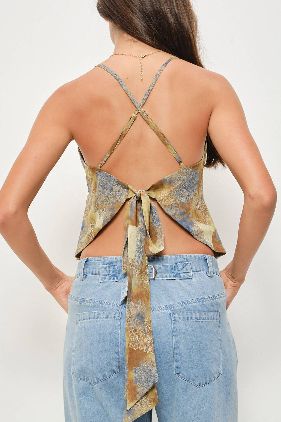 A woman shown from the back wearing a halter-style top with a patterned gold and blue fabric, featuring crisscross straps and a tie-back detail, paired with light-wash wide-leg jeans against a plain white studio background.