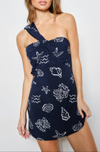 Woman wearing a navy blue one shoulder dress with white ocean sea-themed shell patterns on a plain background