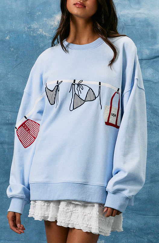 Woman wearing a light blue sweatshirt with graphic designs, white skirt, and white cowboy boots against a textured blue wall.