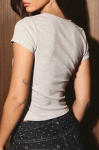 A woman wearing a light gray short-sleeve t-shirt and a dark patterned skirt, photographed from the back against a dark wood-paneled wall.