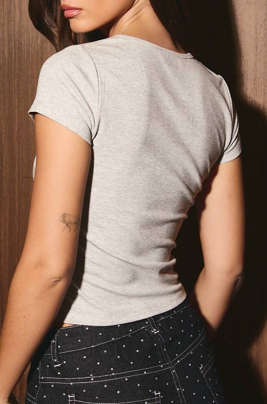 A woman wearing a light gray short-sleeve t-shirt and a dark patterned skirt, photographed from the back against a dark wood-paneled wall.