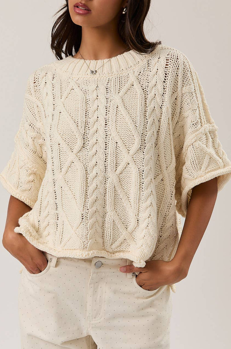 Woman wearing a cream-colored knitted short sleeve sweater against a plain background