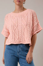 Woman wearing a pink cable knit sweater and blue jeans against a plain background