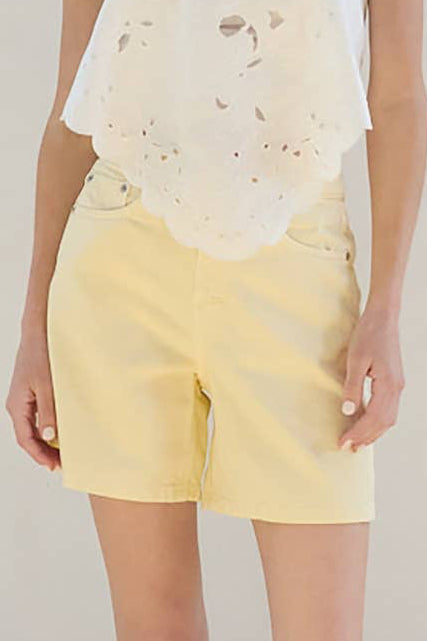 Woman wearing a white top and yellow shorts standing in a room with white walls and wooden floor.