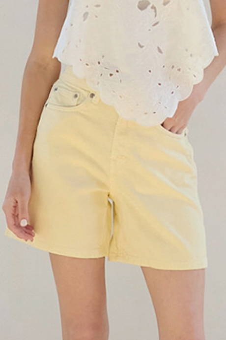 Woman wearing a white top and yellow shorts in a room with a white wall and wooden floor.