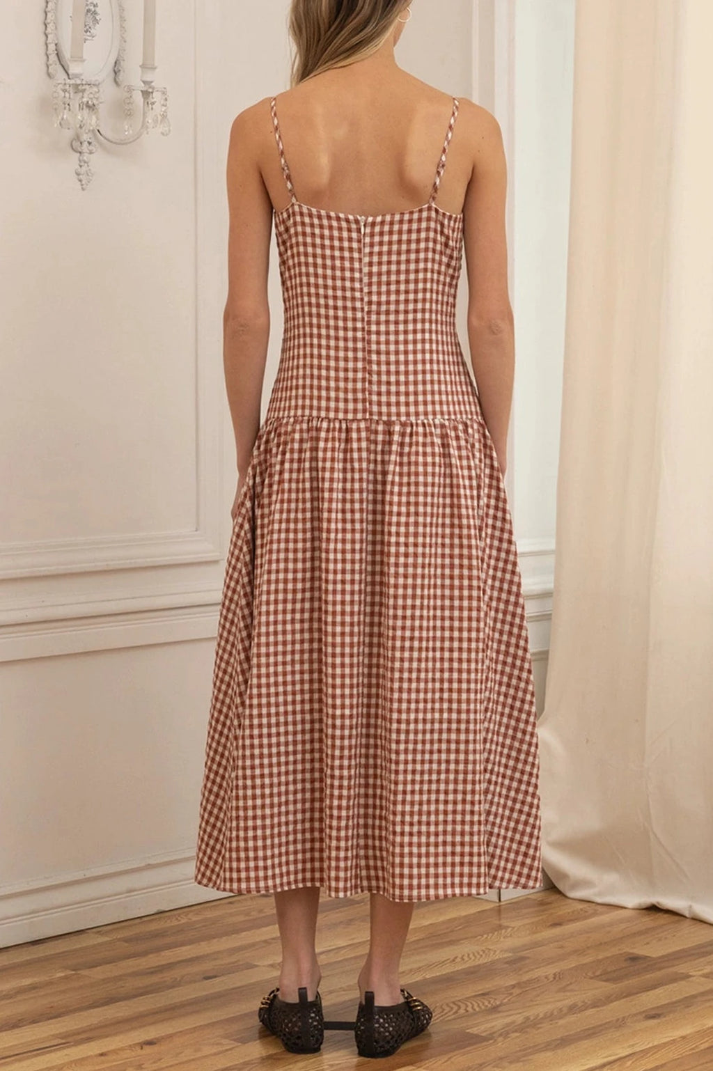Woman wearing a checkered dress standing in a room with wooden floor and white walls.