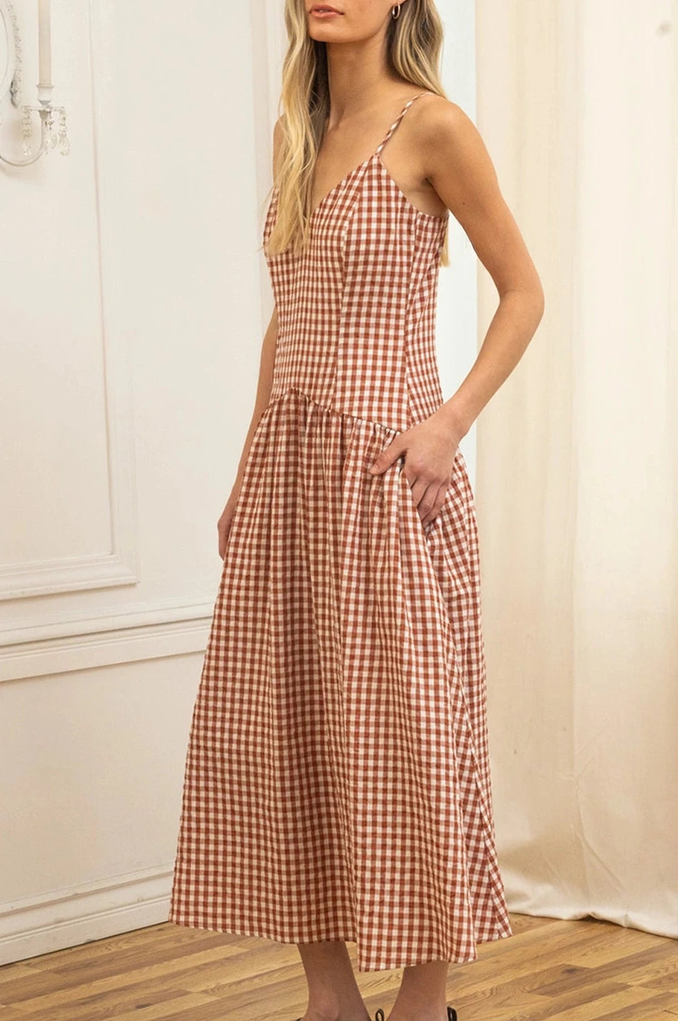 Woman wearing a checkered dress standing in a room with white walls and wooden floor.