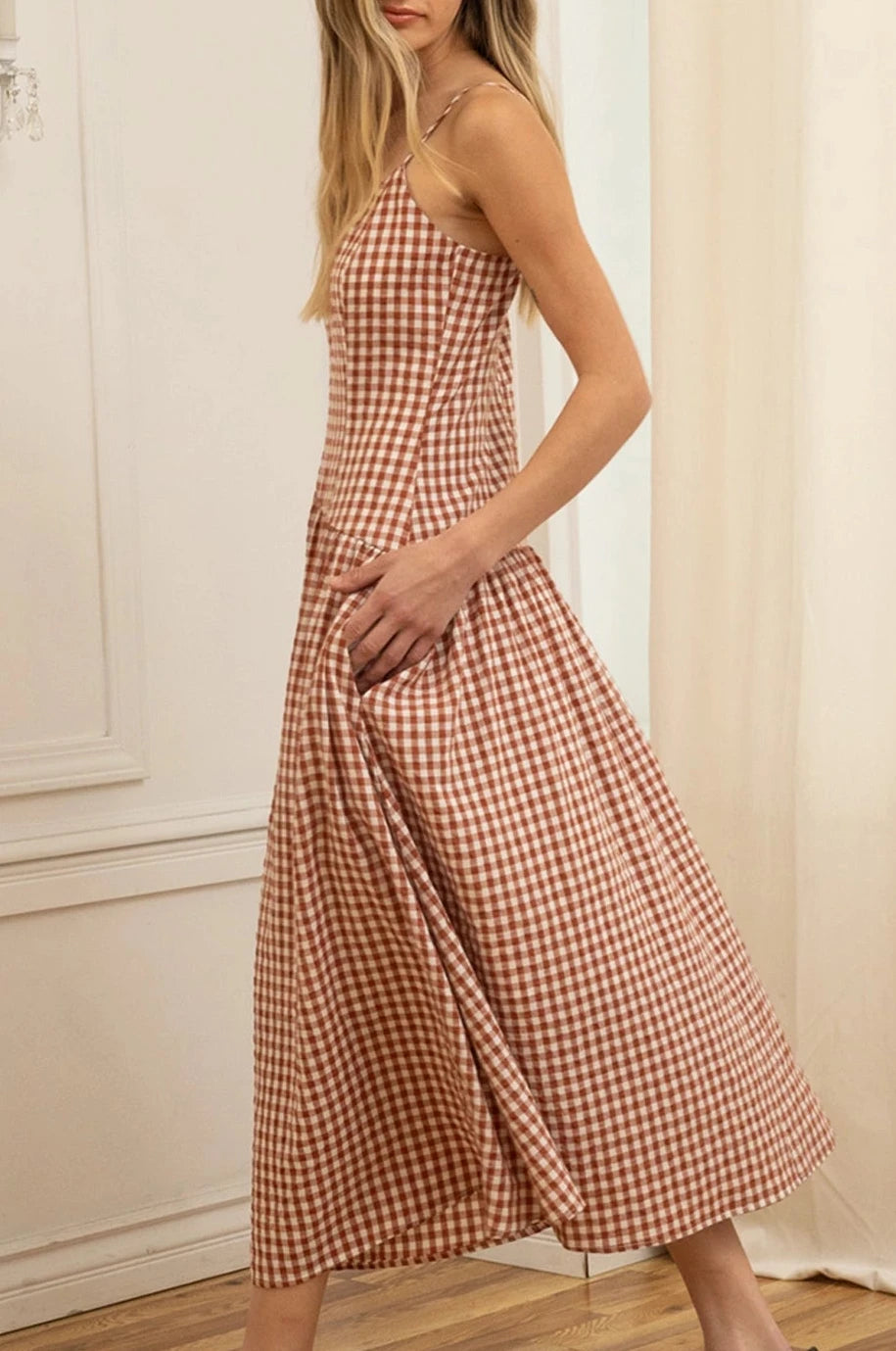 Woman wearing a rust and white checkered dress standing in a room with white walls and wooden floor.
