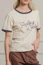 Woman wearing a beige t-shirt with text and brown pants against a white paneled wall.