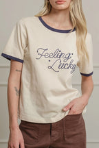 Woman wearing a cream t-shirt with 'Feeling Lucky' text and brown pants against a neutral background