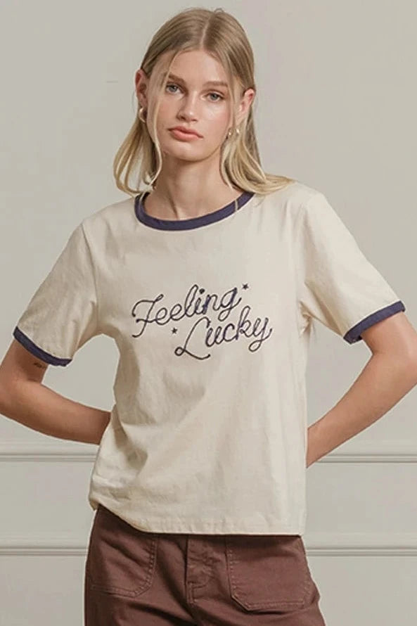 Woman wearing a beige t-shirt with text and brown pants against a neutral wall.