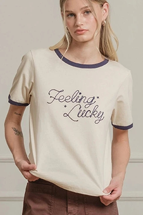 Woman wearing a beige t-shirt with text and brown pants against a white paneled wall.