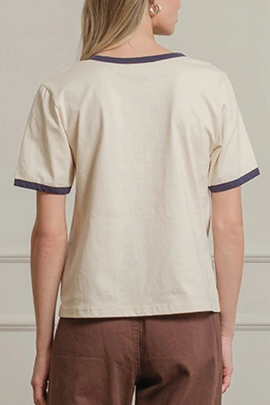 Woman wearing a beige top and brown pants standing against a white paneled wall.