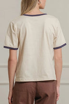 Woman wearing a beige top and brown pants standing against a white paneled wall.