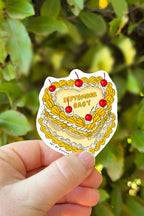 Sticker with a cake design and 'September Baby' text held in front of green foliage