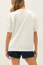 A woman shown from the back wearing a white short-sleeve t-shirt, navy blue shorts, and brown boat shoes against a plain white studio background.