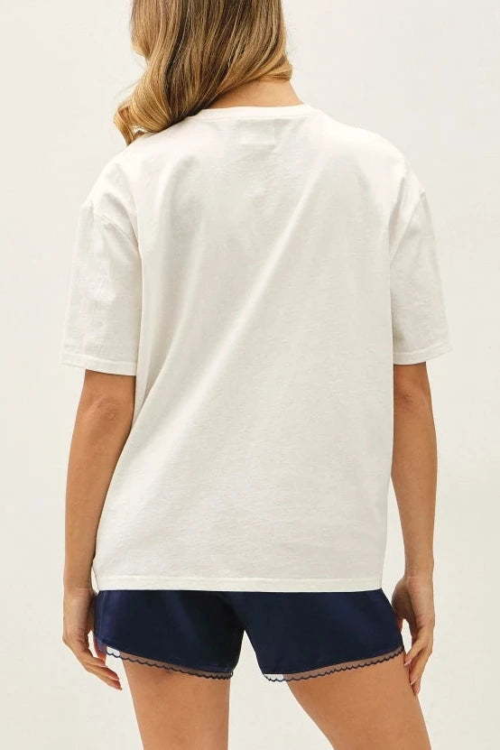 A woman shown from the back wearing a white short-sleeve t-shirt, navy blue shorts, and brown boat shoes against a plain white studio background.